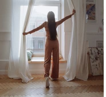 Pijamalarıyla bir kadın yüksek tavanlı yatak odasındaki tül perdeyi açıyor