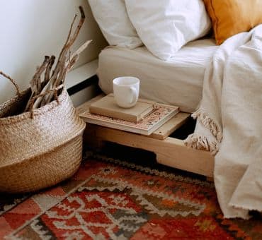 Yatağın üzerinde rahat çarşaf ve pikeler, yanda kitaplar ve yerde el dokuma kilim